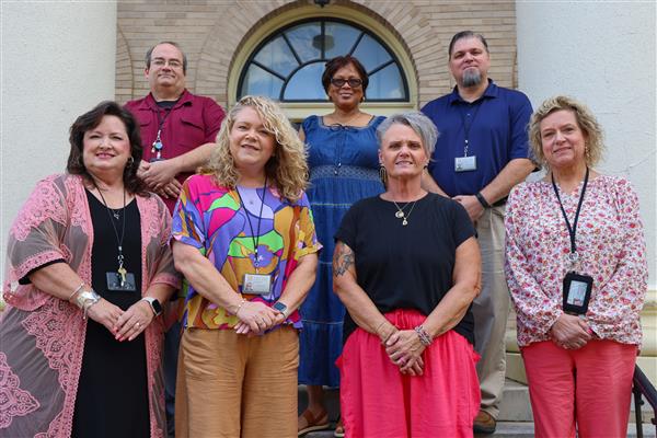 Seven employees stand on the steps outside of Johnson Hall and smile at the camera.